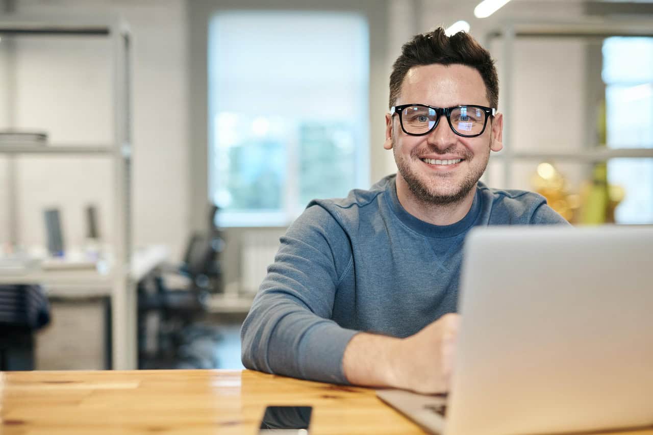 Smiling man wearing glasses using a laptop in an office environment.