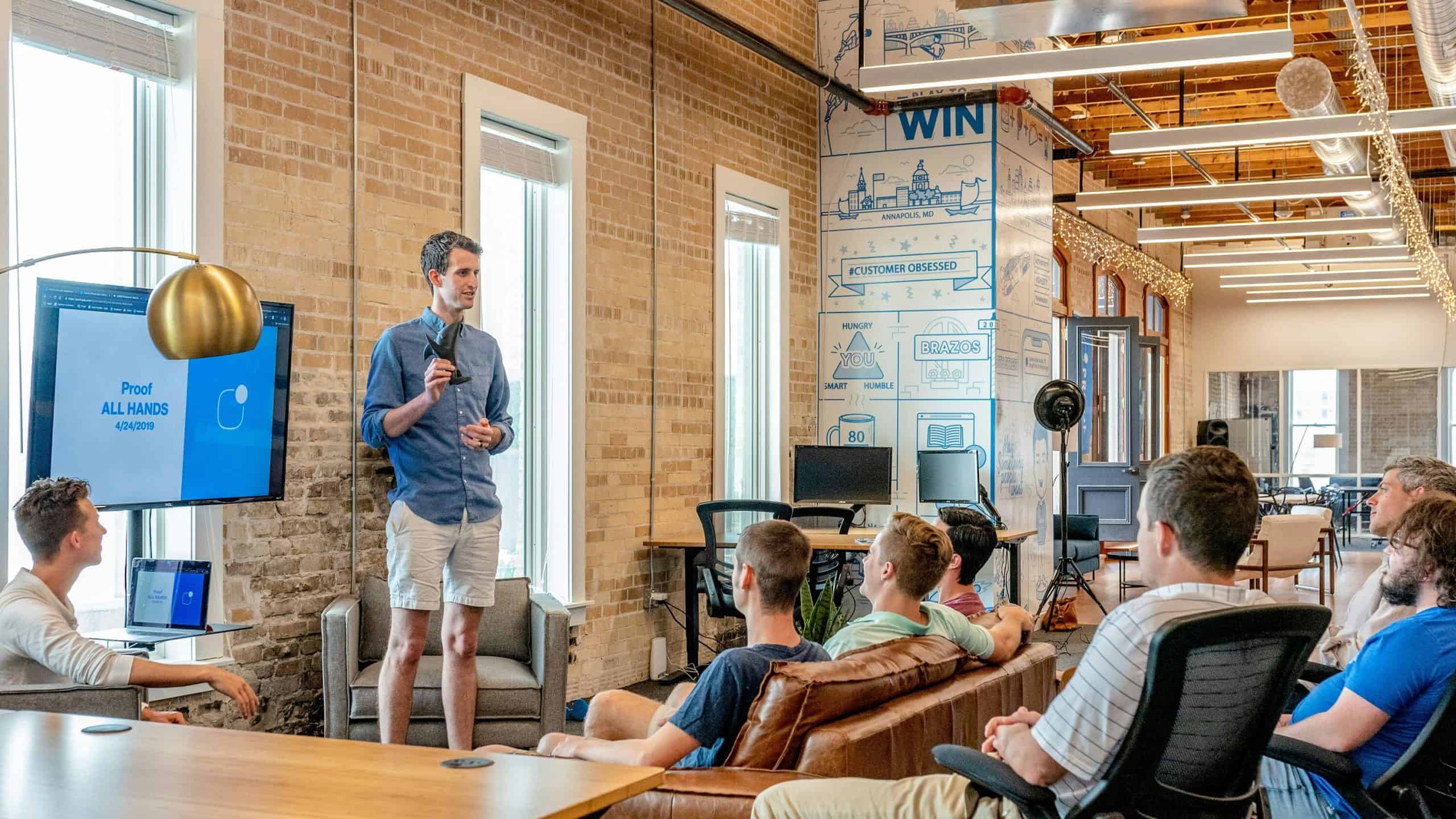 ALT Text: Business team in a modern office listening to a presenter with live chat monitoring screens visible.