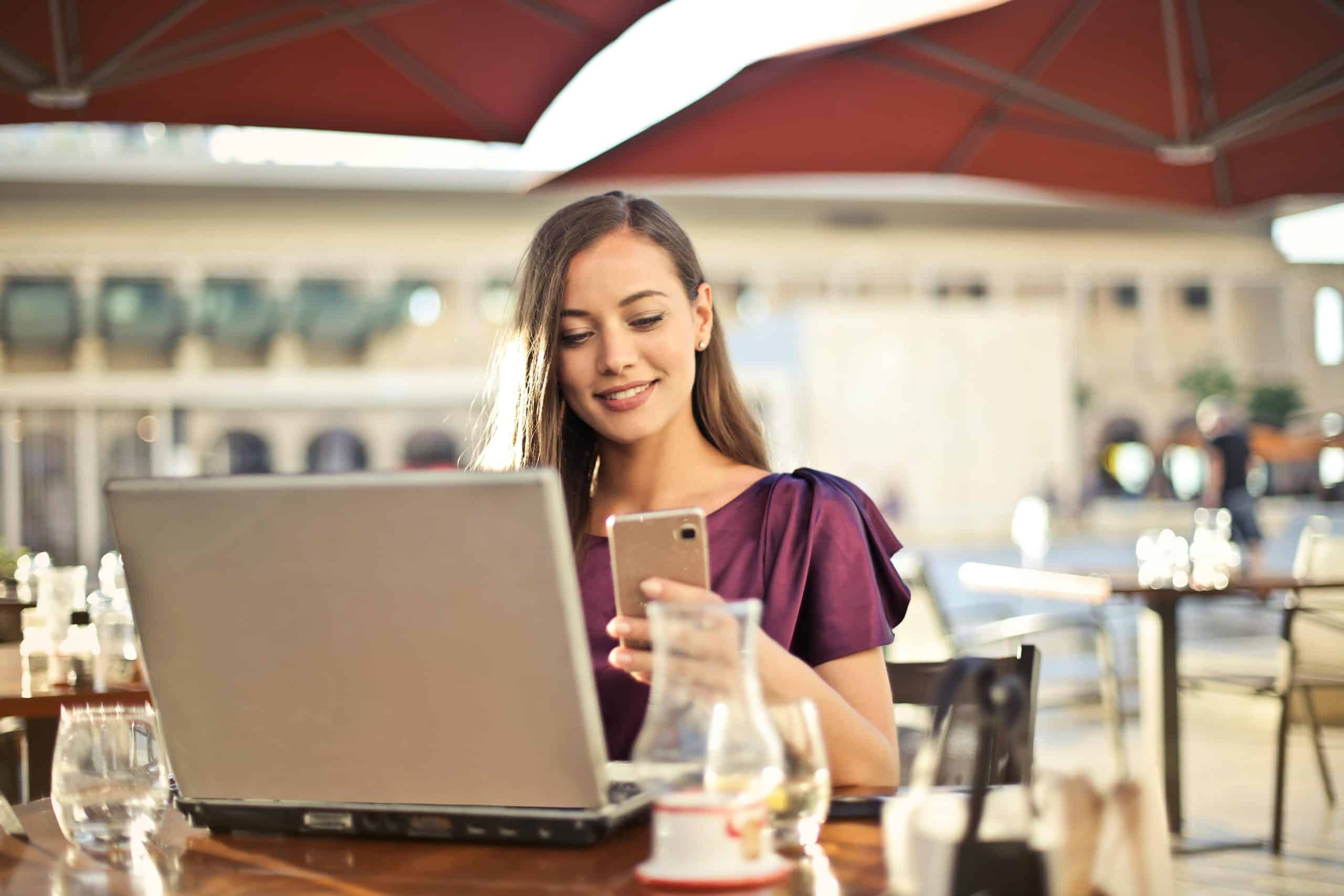 Young woman using smartphone and laptop at outdoor café for live chat support.