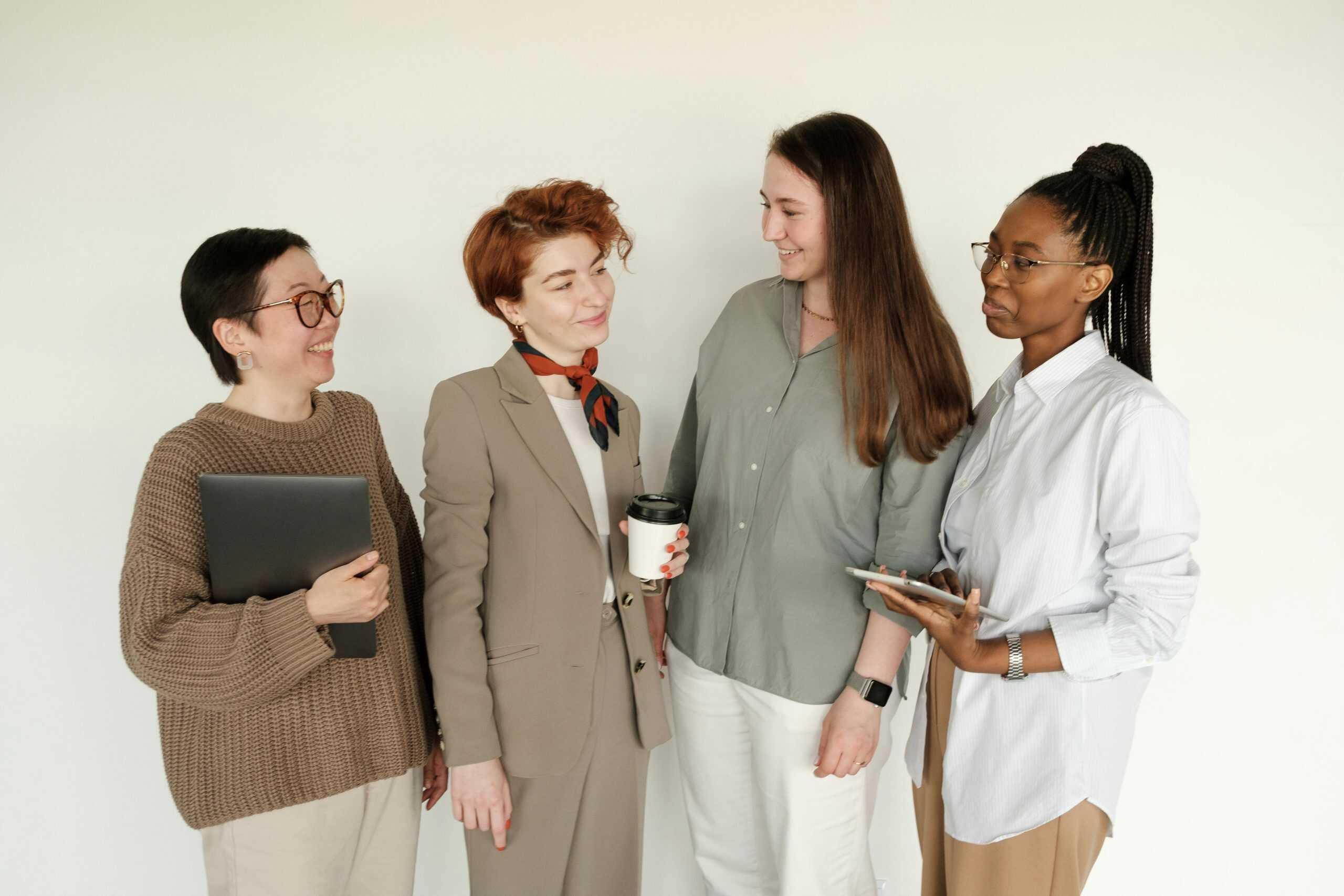 Four colleagues engaged in friendly workplace discussion, with one holding a tablet and another carrying a laptop.