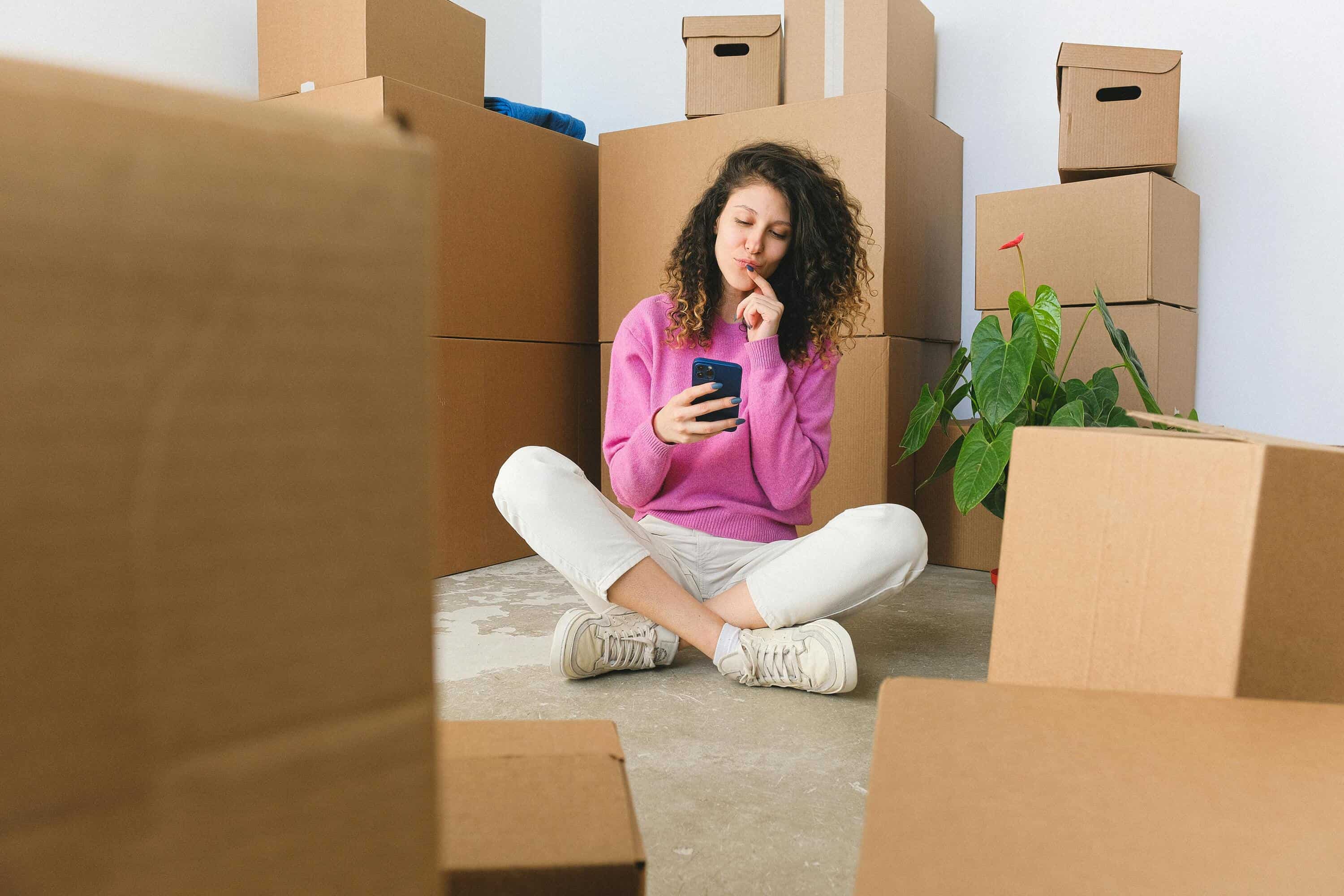 Woman using smartphone among moving boxes for live chat monitoring.